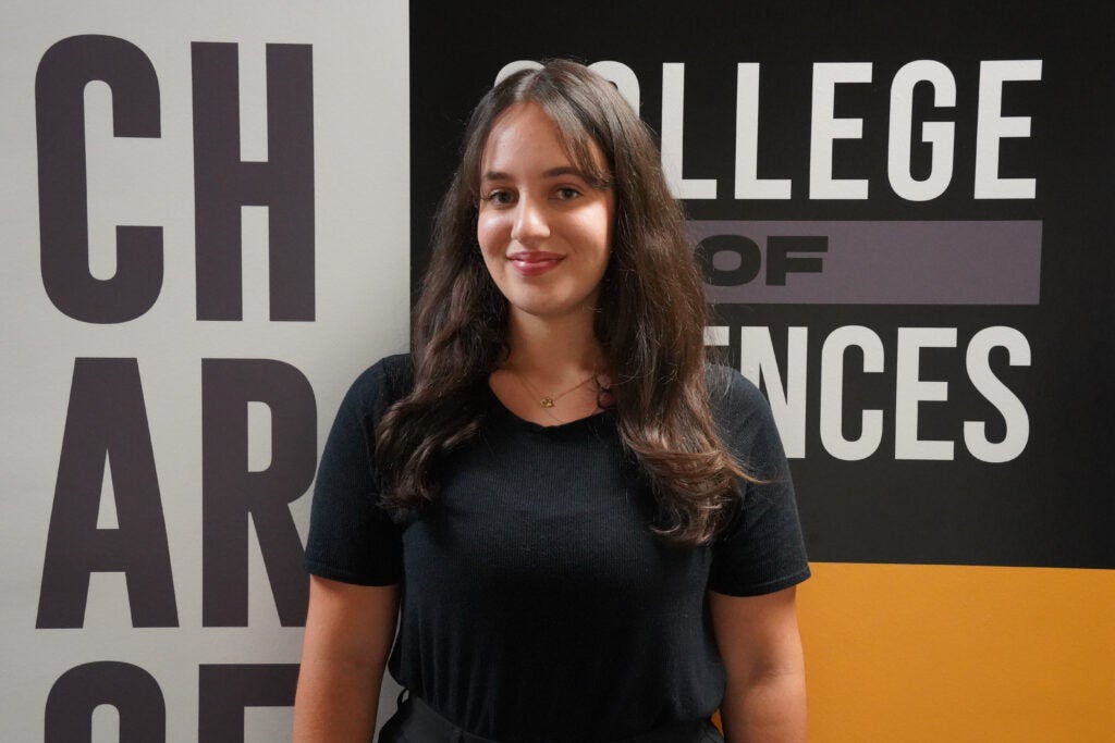 A woman with long brown hair stands in front of a wall with the words "COLLEGE OF SCIENCES" and large partial letters visible in the background.