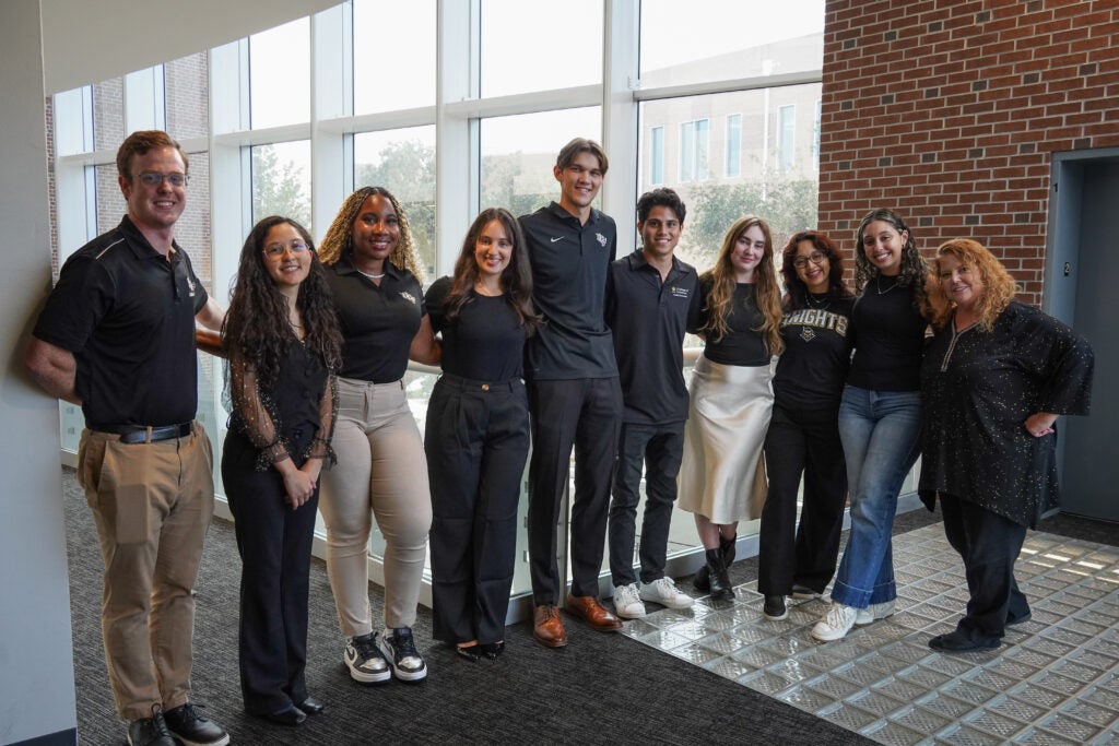 Ten people stand side by side indoors in front of large windows, posing and smiling for the camera.