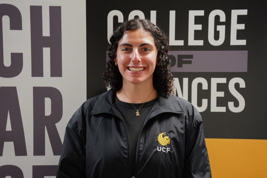 A person with curly hair wearing a black UCF jacket stands smiling in front of a wall with "College of Sciences" written on it.