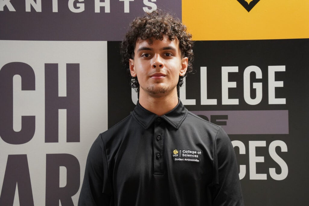 A young man with curly hair wearing a black "College of Sciences Student Ambassador" polo shirt stands in front of a sign with large text and logos.
