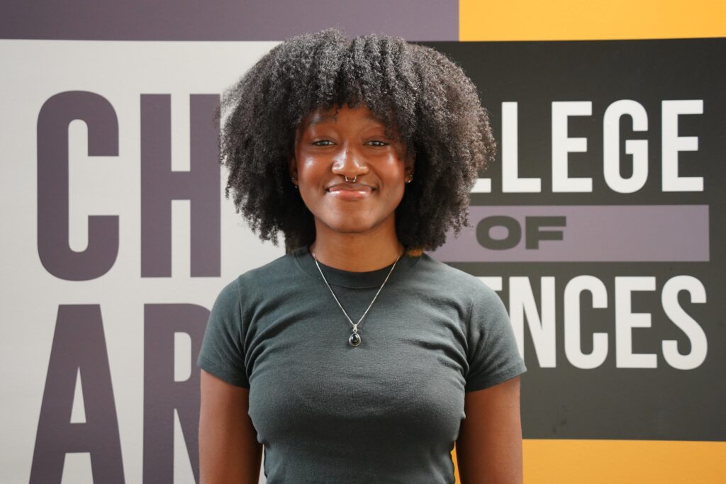 A person with curly hair wearing a dark t-shirt stands in front of a sign that reads "College of Arts and Sciences.