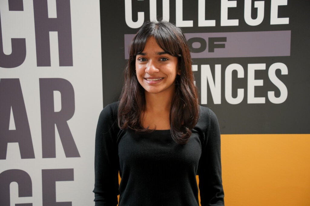 A young woman with long dark hair and a black top stands smiling in front of a sign that reads "College of Sciences.