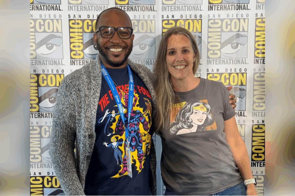 Two people stand smiling in front of a San Diego Comic-Con International backdrop, both wearing graphic T-shirts with comic book characters.
