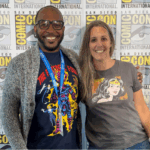 Two people stand smiling in front of a San Diego Comic-Con International backdrop, both wearing graphic T-shirts with comic book characters.