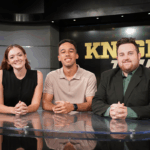 Three people sit behind a news desk with the words "KNIGHT TIME" displayed on a screen behind them. They are facing the camera and smiling.