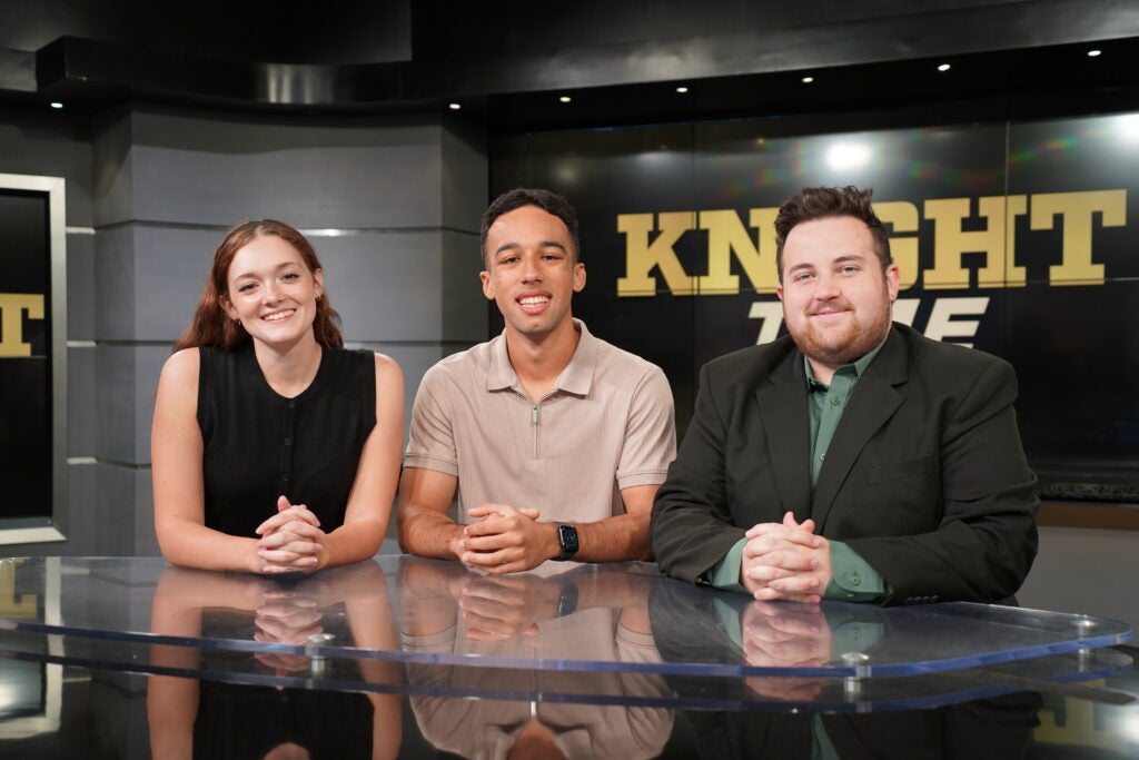 Three people are seated at a news desk in a studio with "KNIGHT" visible on the screen behind them. They are smiling and facing the camera.
