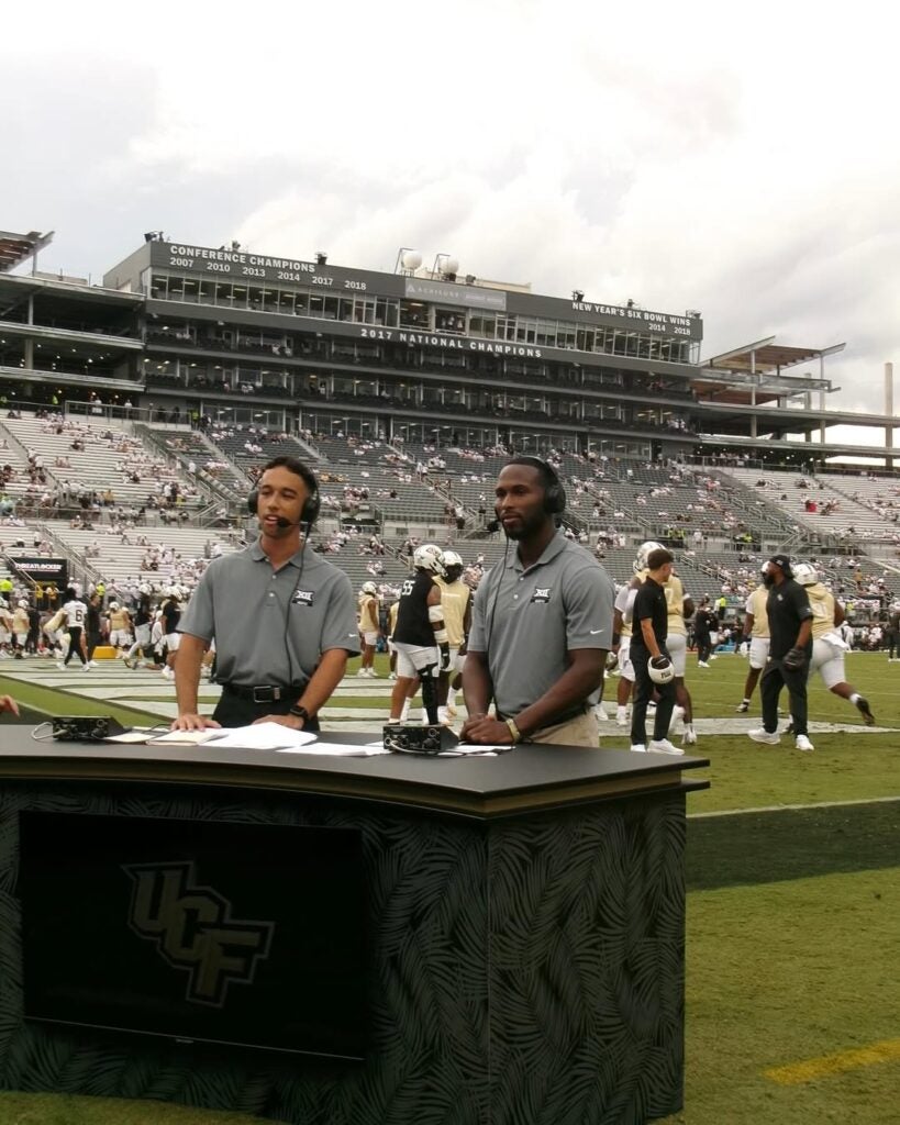 Two sports commentators stand at a desk on a football field, with players practicing and a stadium with fans in the background.