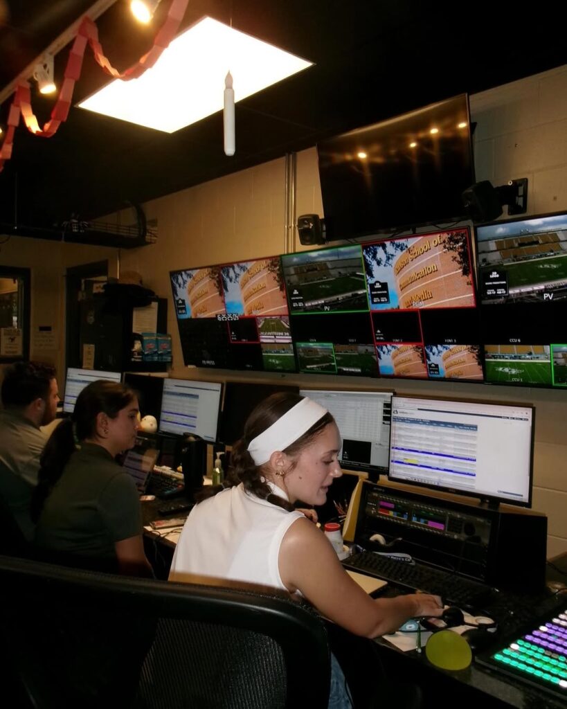Three people work at a control room desk with multiple monitors displaying video feeds, computers, and control panels.