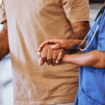 A nurse in blue scrubs holds the hand of an older man with a cane, offering support and care.