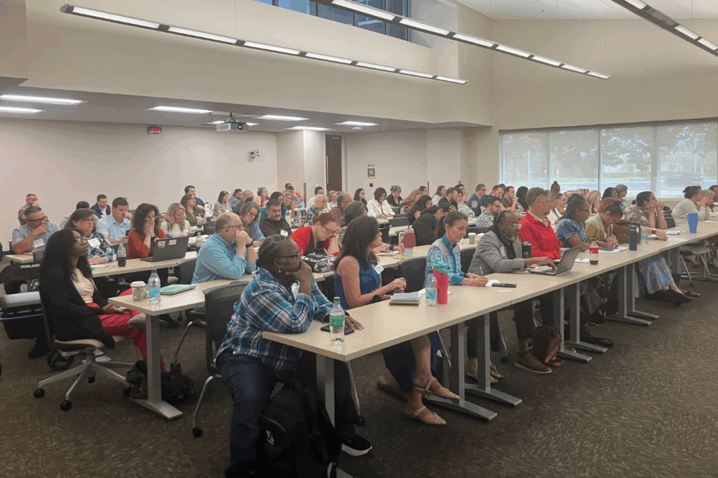 A large group of adults sit at tables in a well-lit classroom, attentively facing the front, with notebooks, laptops, and drinks on the tables.