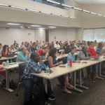 A large group of adults sit at tables in a well-lit classroom, attentively facing the front, with notebooks, laptops, and drinks on the tables.