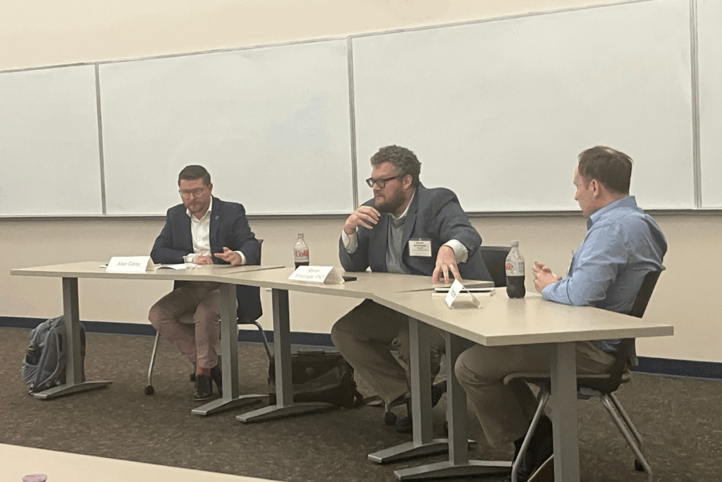 Three men sit at desks in a classroom, engaged in discussion. Two have soda bottles in front of them. A blank whiteboard is in the background.