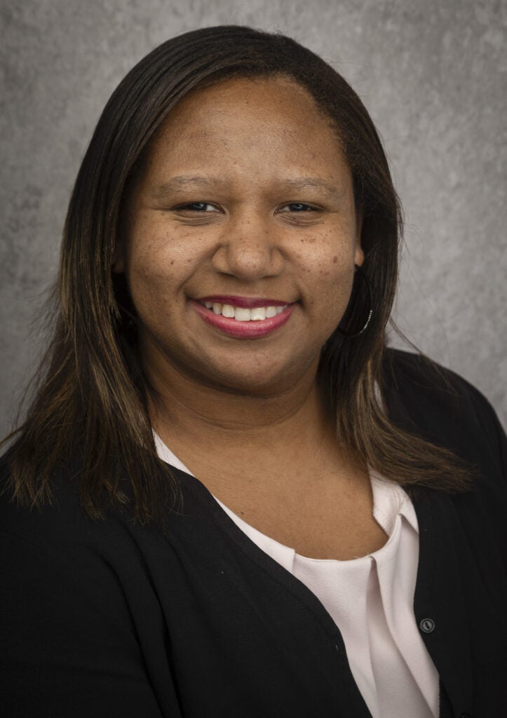 A woman with straight brown hair, wearing a black blazer over a light blouse, smiles at the camera against a gray textured background.