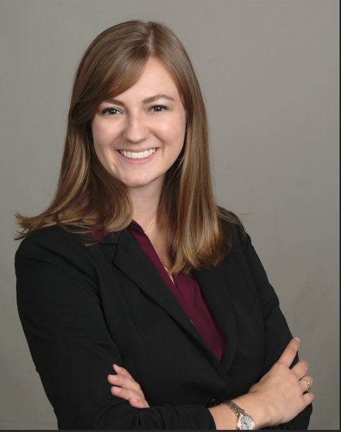 A woman with straight, shoulder-length light brown hair poses smiling with arms crossed, wearing a black blazer over a burgundy top against a plain gray background.