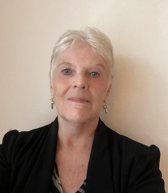 An older woman with short white hair, wearing earrings and a black blazer, poses in front of a plain light-colored wall.