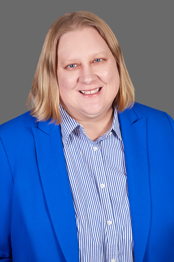 Person with light hair wearing a blue blazer and striped shirt, smiling in front of a plain gray background.
