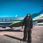 A person wearing sunglasses stands on an airport tarmac next to a small private jet with blue and green stripes.