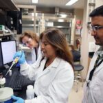 Three scientists in lab coats work together in a laboratory; one uses a pipette while the others observe and monitor data on computer screens.