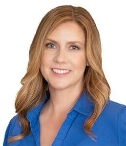 A woman with long, wavy blonde hair and light eyes is smiling. She is wearing a blue collared shirt and is pictured against a plain white background.