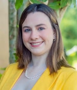 A woman with straight brown hair, wearing a yellow blazer, a pearl necklace, and small hoop earrings, smiles outdoors in front of green foliage.