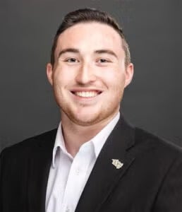A man in a black suit jacket and white shirt smiles at the camera against a plain dark background.