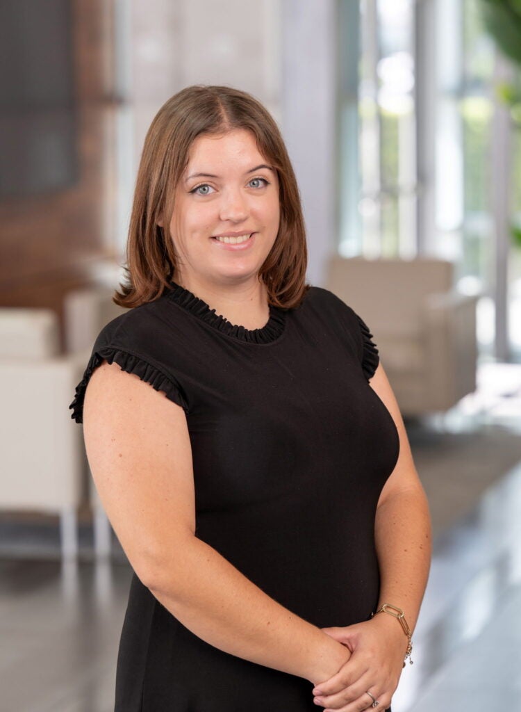 A woman with shoulder-length brown hair wearing a black dress stands indoors with her hands clasped, smiling at the camera. The background includes neutral-colored chairs and large windows.