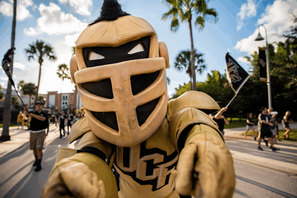 A person in a gold and black knight mascot costume participates in an outdoor parade, with people and UCF flags visible in the background.