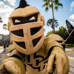 A person in a gold and black knight mascot costume participates in an outdoor parade, with people and UCF flags visible in the background.