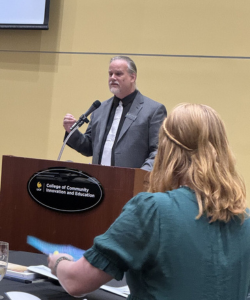 A man in a suit speaks at a podium labeled "College of Community Innovation and Education" as a woman listens in the foreground.