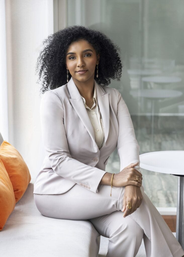 A woman in a light-colored suit sits on a bench next to orange pillows, with her hands resting on her knee, in a modern, bright room.