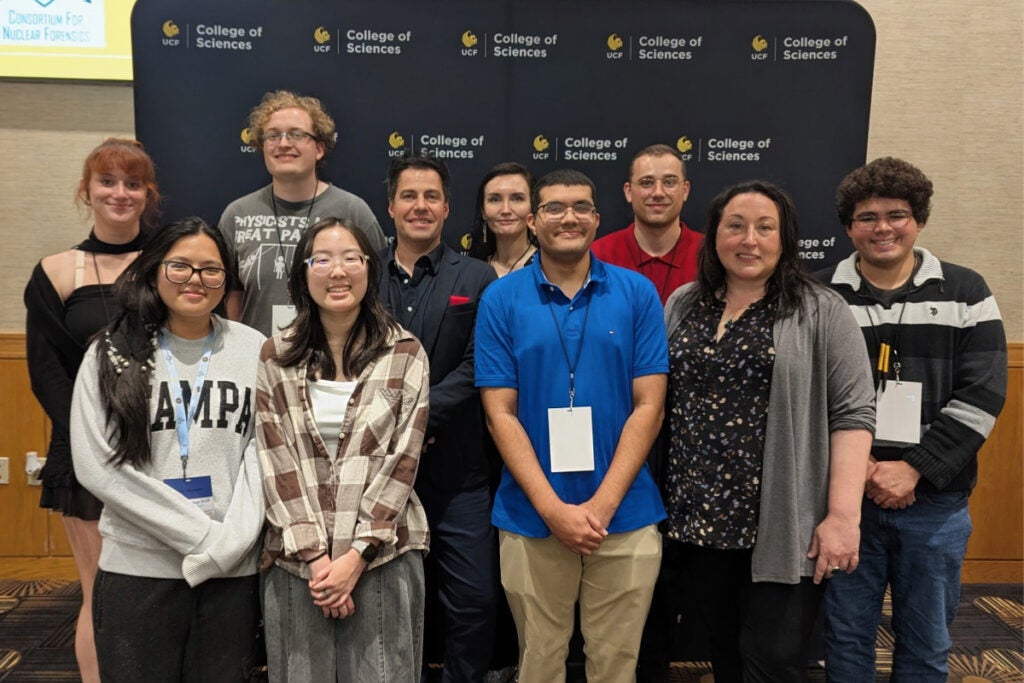 A group of eleven people pose for a photo indoors in front of a College of Sciences backdrop, all wearing name badges.