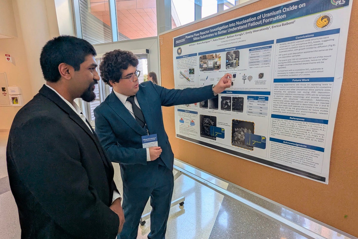Two men in suits stand in front of a scientific poster, with one man pointing at data on the poster while discussing its content with the other.