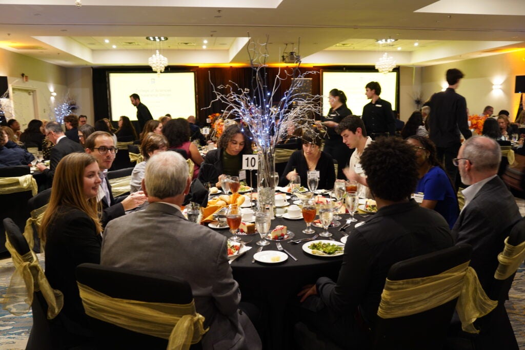 People are seated around a decorated round table at a formal event or banquet, with plates of food, drinks, and table centerpiece visible. Other attendees and servers are in the background.