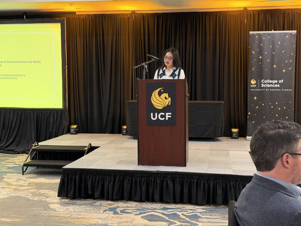 A woman stands at a podium with a UCF logo, speaking at a College of Sciences event. A presentation is displayed on a screen to the left.