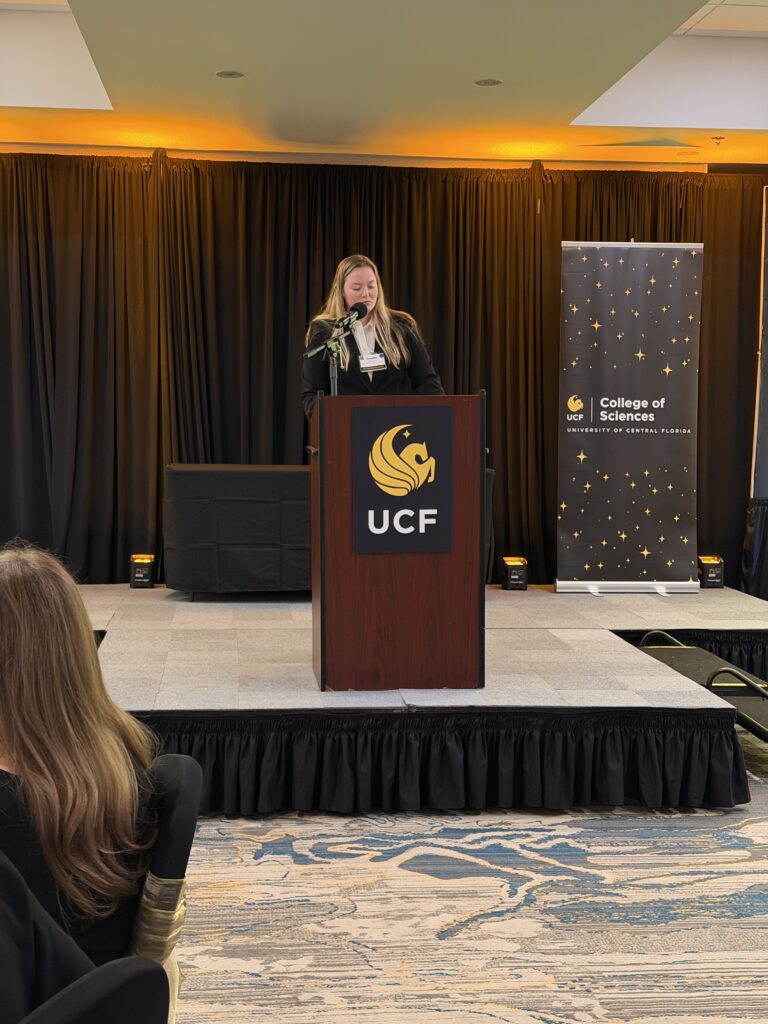 A woman stands at a podium with the UCF logo, delivering a speech at a College of Sciences event, with black and gold decor in the background.