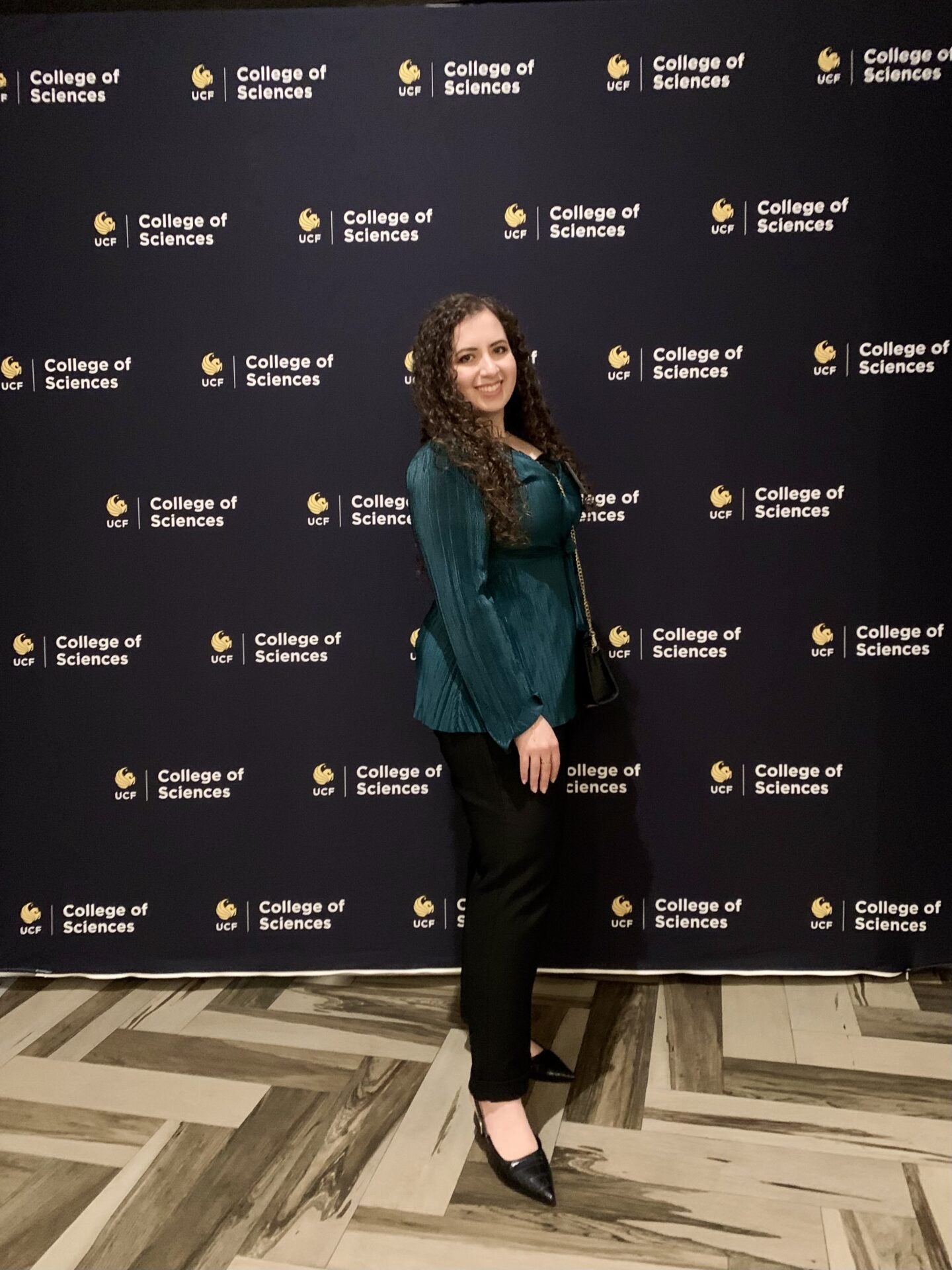 A woman stands in front of a backdrop with "UCF College of Sciences" logos, wearing a green jacket, black pants, and black heels.