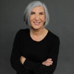 Older woman with gray hair, wearing a black top, stands against a plain gray background with arms crossed and a slight smile.