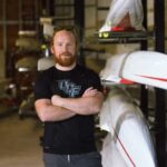 A man with a beard and sunglasses on his head stands with arms crossed in front of stored rowing shells inside a boathouse.