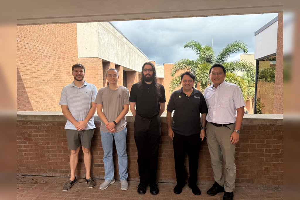 Five men stand in a row outside on a brick terrace with a building and palm tree in the background.