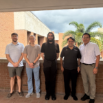 Five men stand in a row outside on a brick terrace with a building and palm tree in the background.