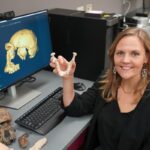A woman in a lab holds a jawbone model beside a computer displaying a 3D skull scan, with various bone models and fossils on the desk.