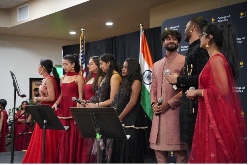 A group of people in traditional Indian attire stand on stage with microphones, in front of U.S. and Indian flags and a "College of Sciences" backdrop.