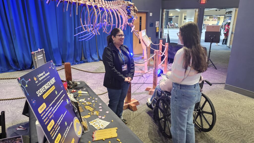 Two women converse at a science exhibit table with informational displays, science-themed items, and a large skeleton model in the background. One woman uses a wheelchair.