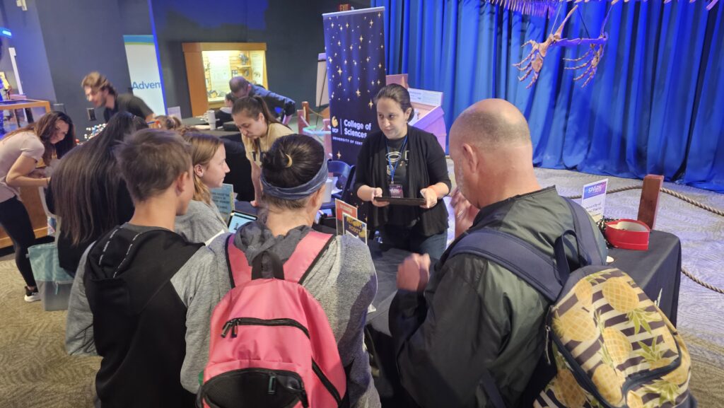 A group of people gathers around a table with science displays, while a presenter explains something to them in an indoor event setting.