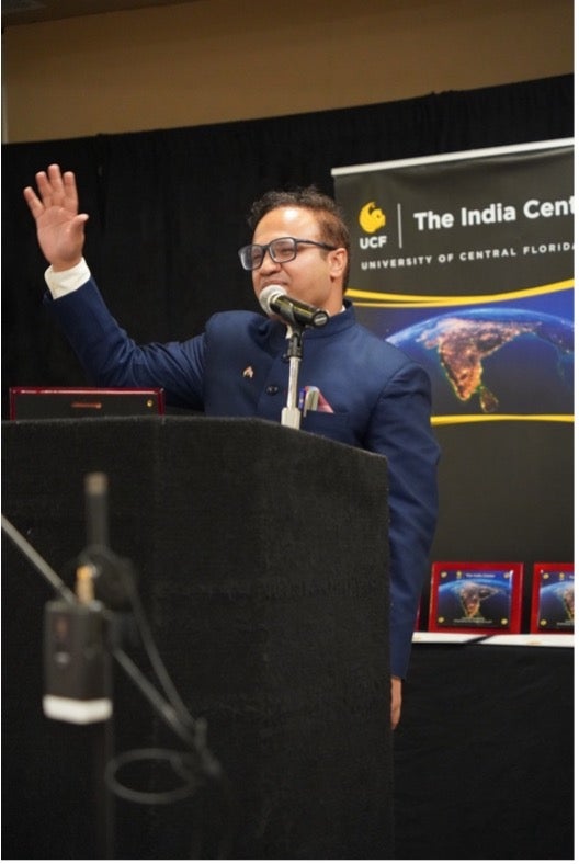 A man in a blue suit speaks at a podium with a raised hand. Behind him is a sign for The India Center at UCF and an image of India from space.
