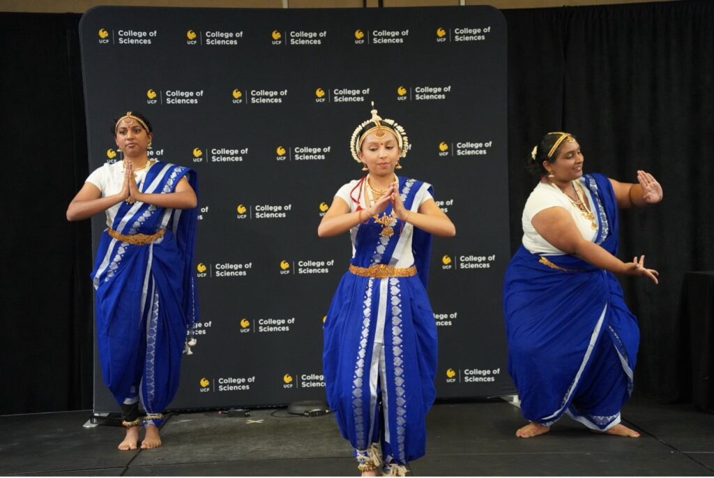 Three dancers in traditional blue and white attire perform on stage in front of a College of Sciences backdrop.
