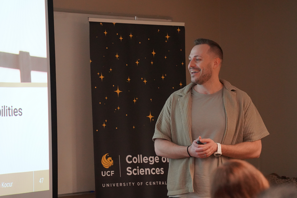 A person stands and smiles while giving a presentation in front of a UCF College of Sciences banner and a projected slide.
