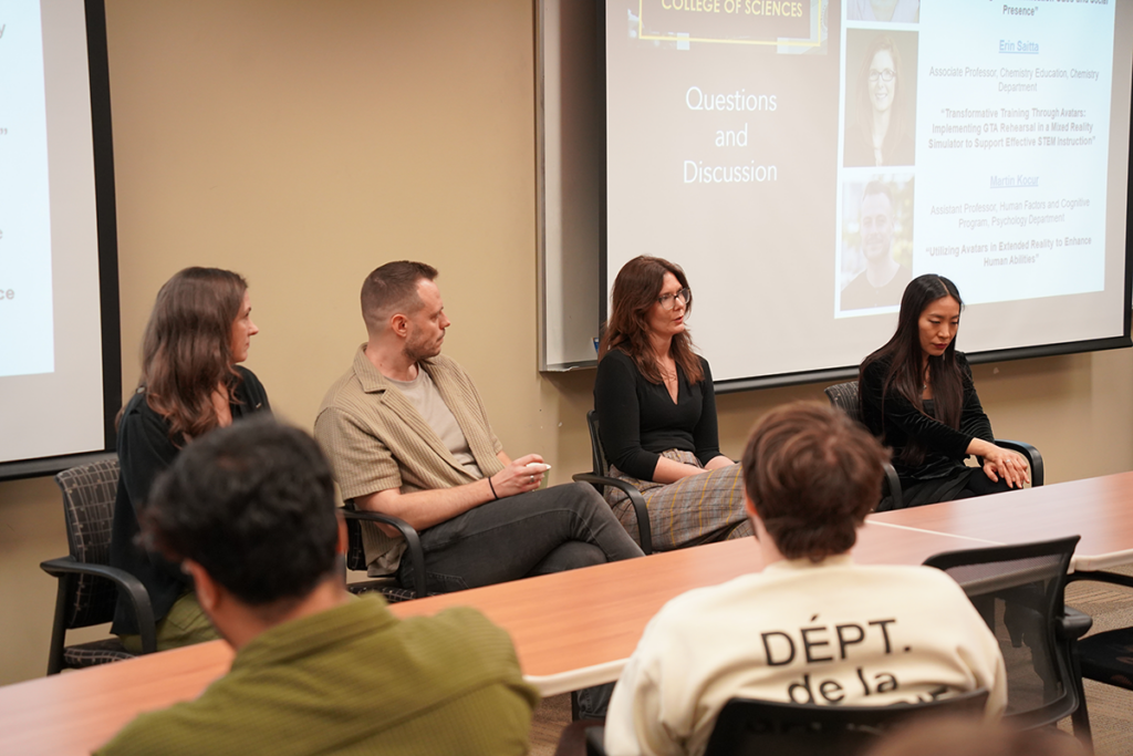 Four panelists sit at the front of a classroom during a questions and discussion session, with slides projected behind them and audience members visible in the foreground.