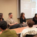 Four panelists sit at the front of a classroom during a questions and discussion session, with slides projected behind them and audience members visible in the foreground.