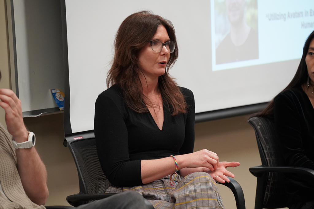 A woman wearing glasses and plaid pants speaks while seated in a panel discussion, gesturing with her hands. A presentation screen is visible in the background.